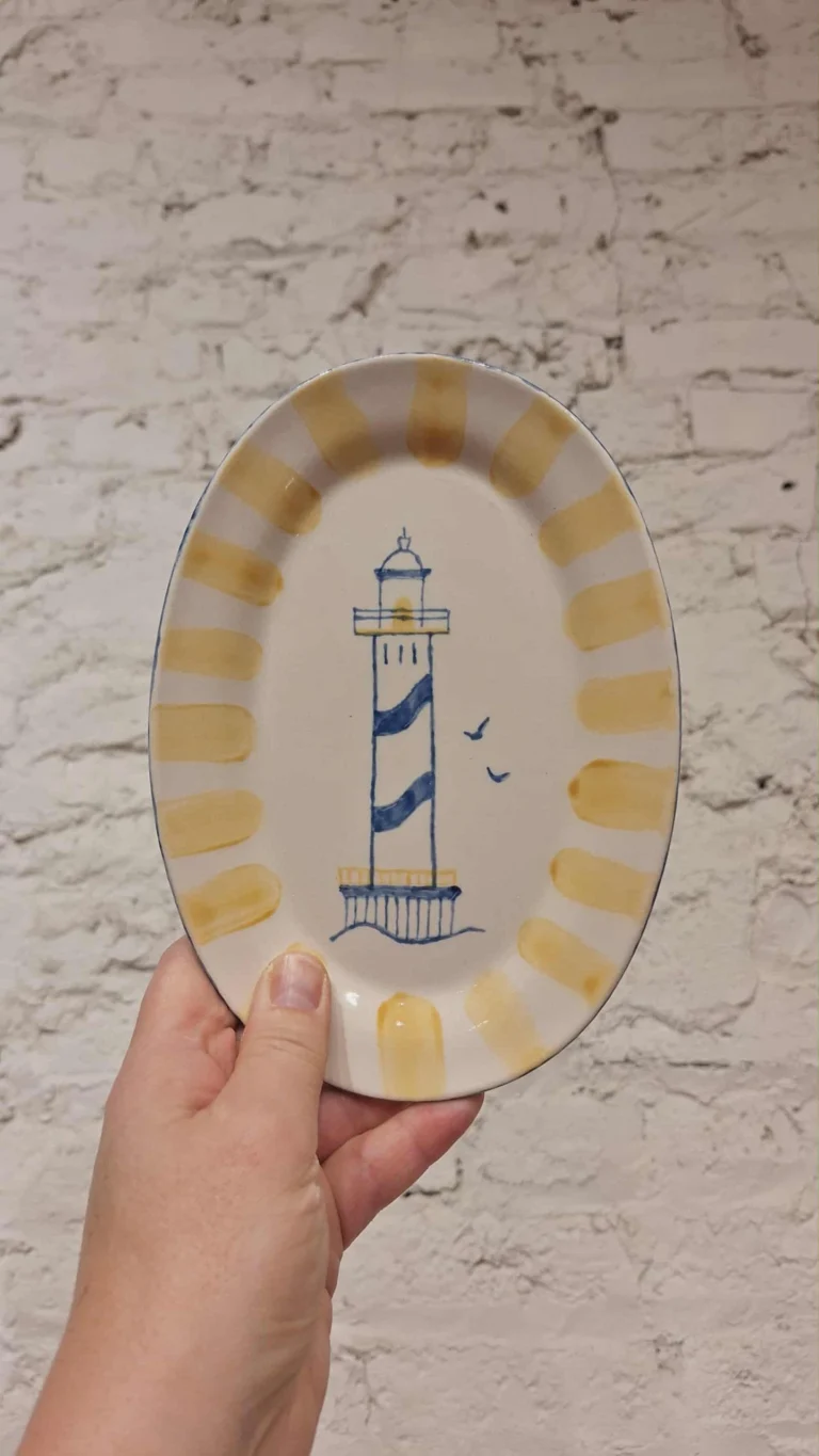 Hand holding an oval plate painted with a blue-striped lighthouse and a yellow border against a whitewashed brick wall
