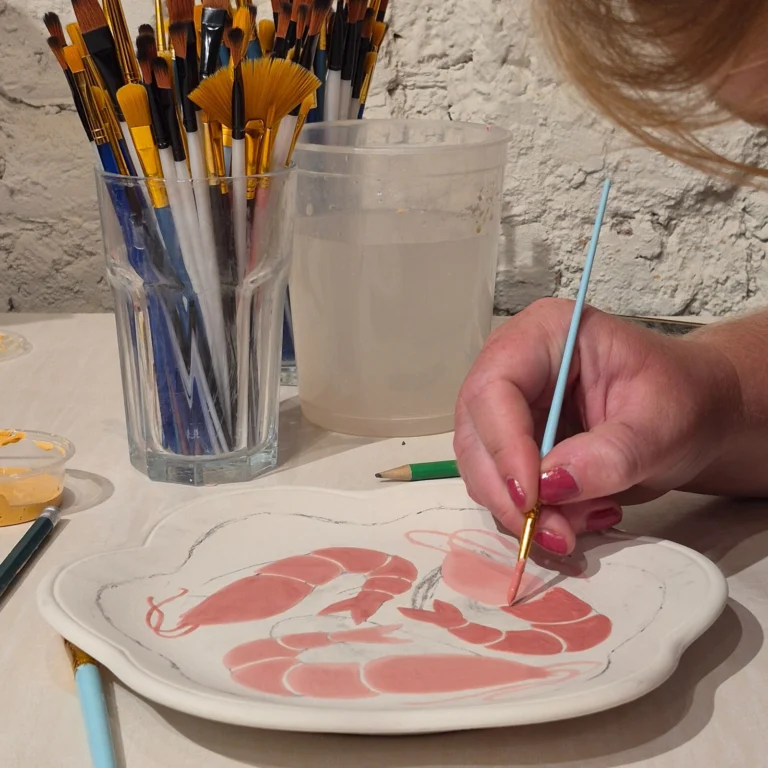 Hand painting pink prawns on a white ceramic plate, with a glass of brushes and a water pot on the table