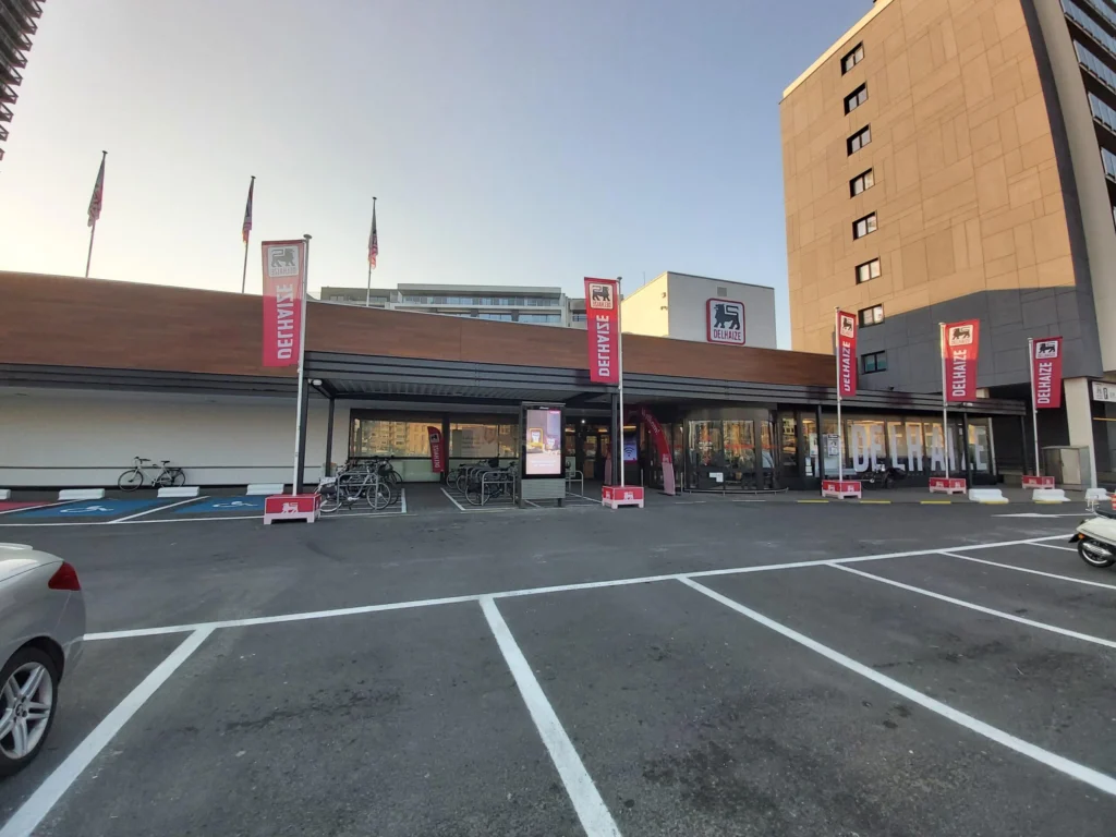 Low supermarket building with red Delhaize flags along the entrance and an empty car park in front
