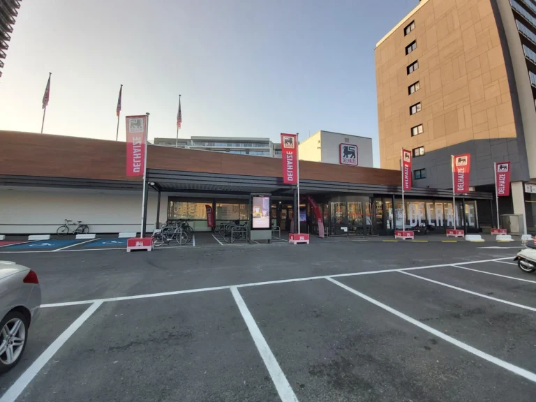 Low supermarket building with red Delhaize flags along the entrance and an empty car park in front