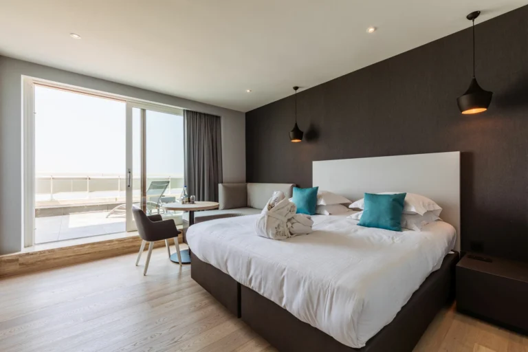 Deluxe room with a double bed, teal cushions, pendant lights and sliding doors to a balcony overlooking the sea