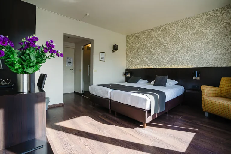 Sunlit double room with twin beds, a grey bed runner, yellow armchair and a vase of purple flowers