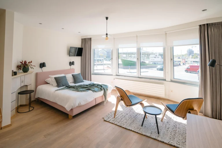 Bright double room with a pink bed, two lounge chairs and wide windows facing the harbour quay