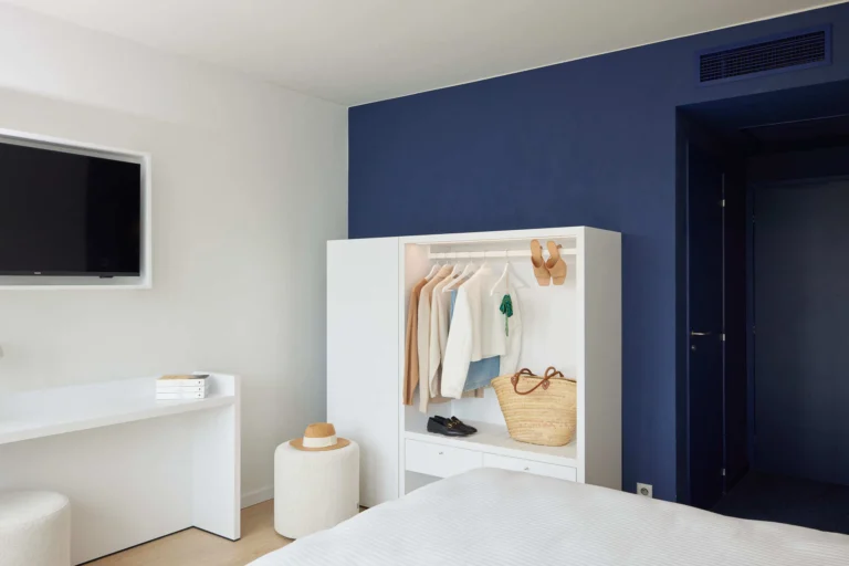 Bright hotel room with a navy feature wall, open white wardrobe, desk and wall-mounted television