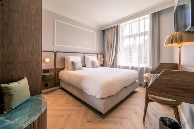 Elegant double room with herringbone floor, upholstered headboard, brass desk lamp and tall windows