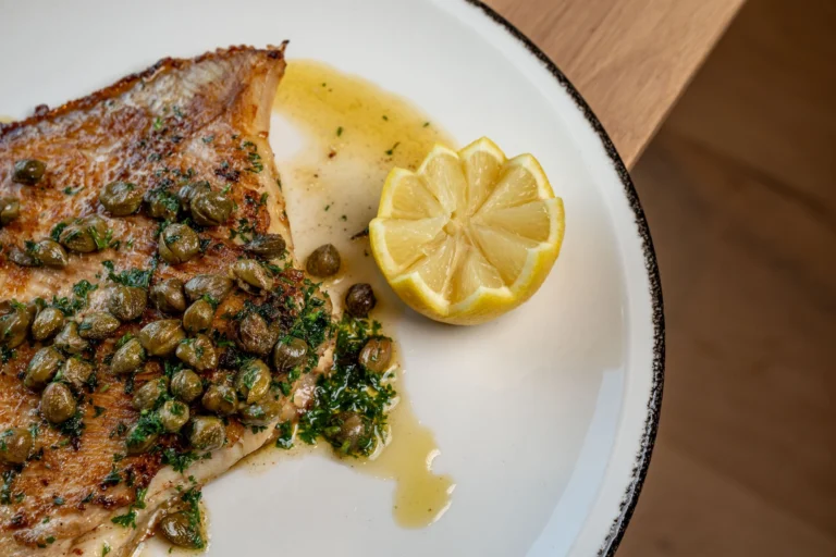 Pan-fried skate wing topped with capers and parsley, with half a lemon, on a white plate