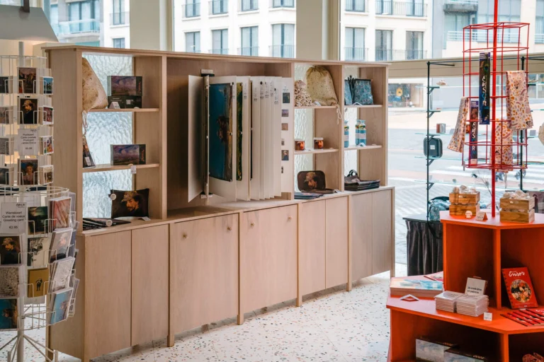 Museum shop with Ensor postcards, books and souvenirs on light wooden shelves by a large window