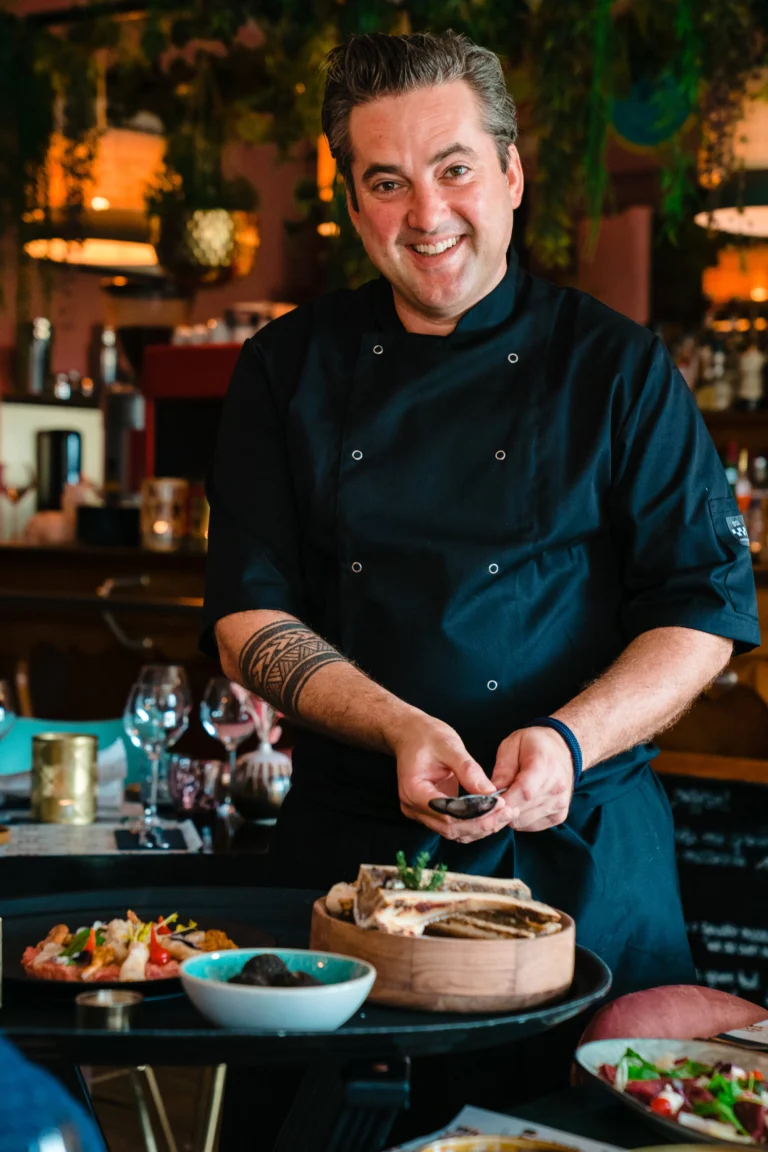 A smiling chef in a black jacket standing at a counter with plates of food in front of him, inside the restaurant