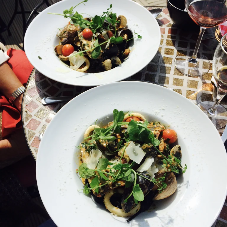 Two white bowls of seafood pasta with mussels, squid, cherry tomatoes and herbs on a mosaic-topped table, beside glasses of wine