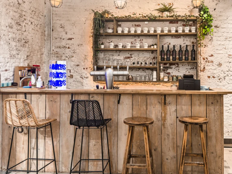 Small bar with a reclaimed-wood counter, rattan and wooden bar stools, a beer tap and shelves of mugs, glasses and wine bottles against a whitewashed brick wall
