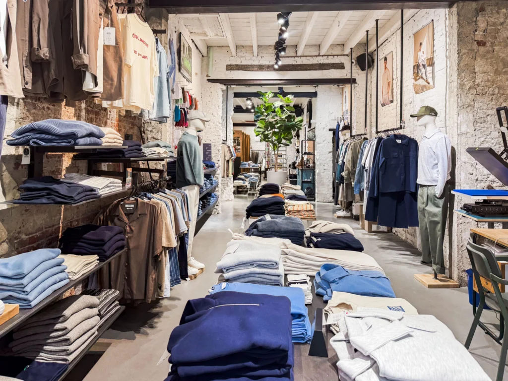 Long menswear shop with whitewashed brick walls and beams, tables of folded knitwear and sweatshirts, shirts on wall rails, mannequins and a tall fig plant at the back