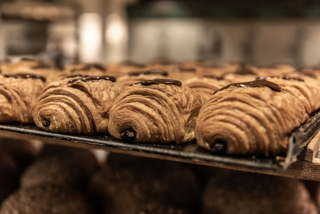 Rows of chocolate croissants on a baking tray, with bread rolls out of focus behind