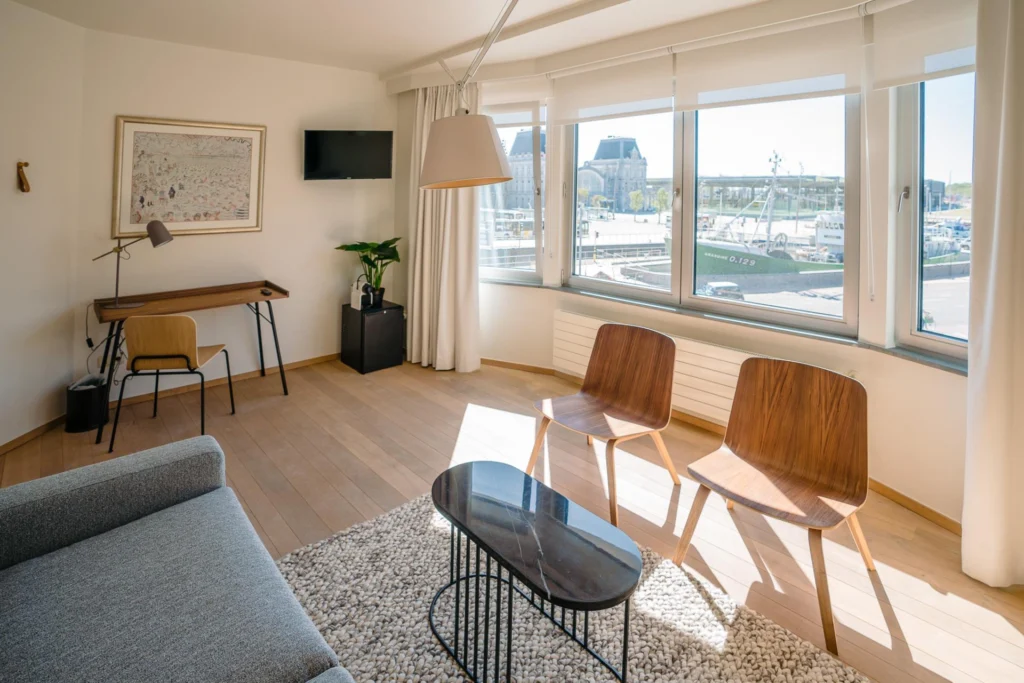 Sunny lounge with a grey sofa and wooden chairs, its bay windows overlooking fishing boats and the harbour