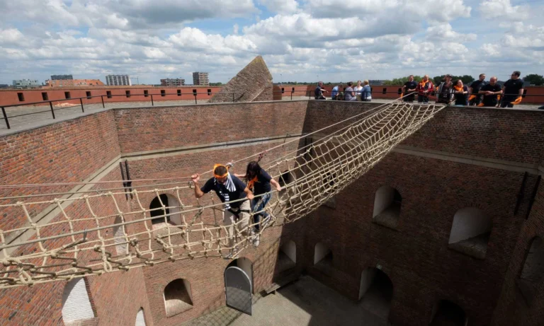 Two people crossing a rope net bridge over the fort's brick inner courtyard while a group watches from the ramparts