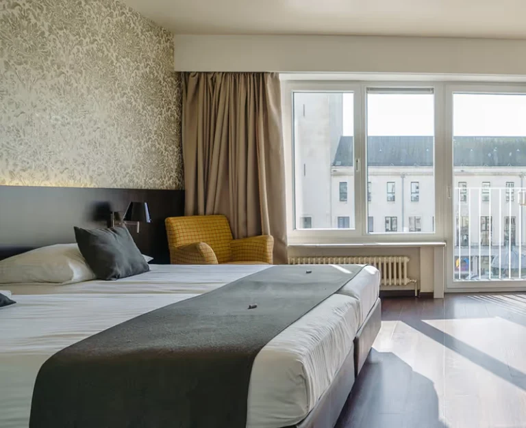 Front-facing hotel room with a double bed and large windows looking onto buildings across the square