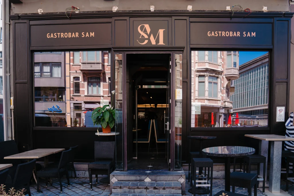 Dark frontage of Gastrobar Sam with large windows, an SM monogram above the door and tables outside
