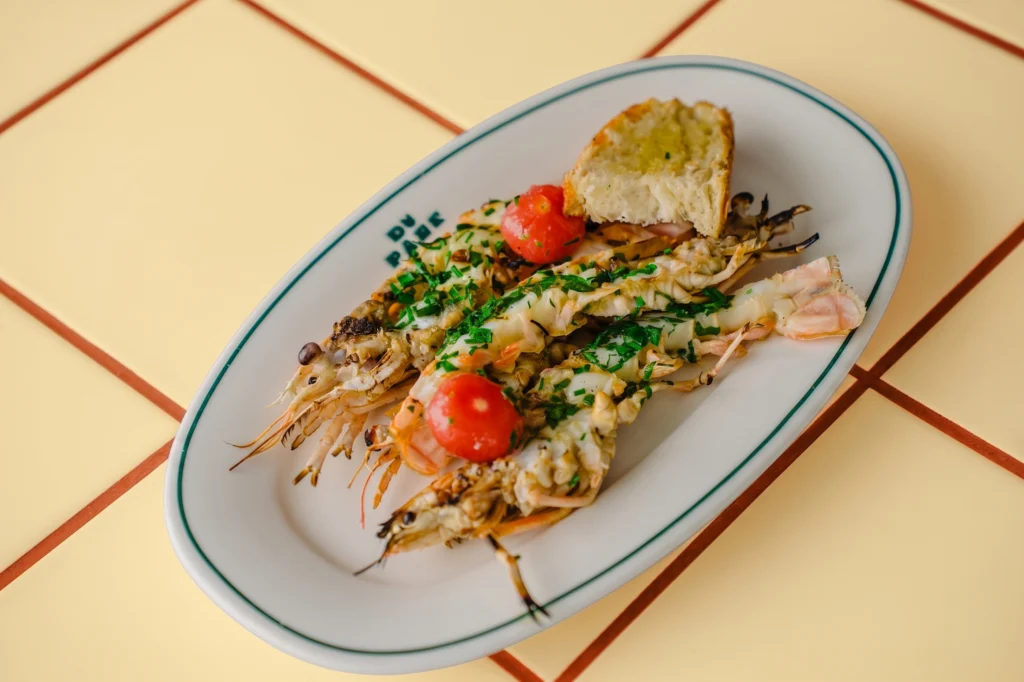 Grilled langoustines with herbs, cherry tomatoes and toasted bread on an oval plate on yellow tiles