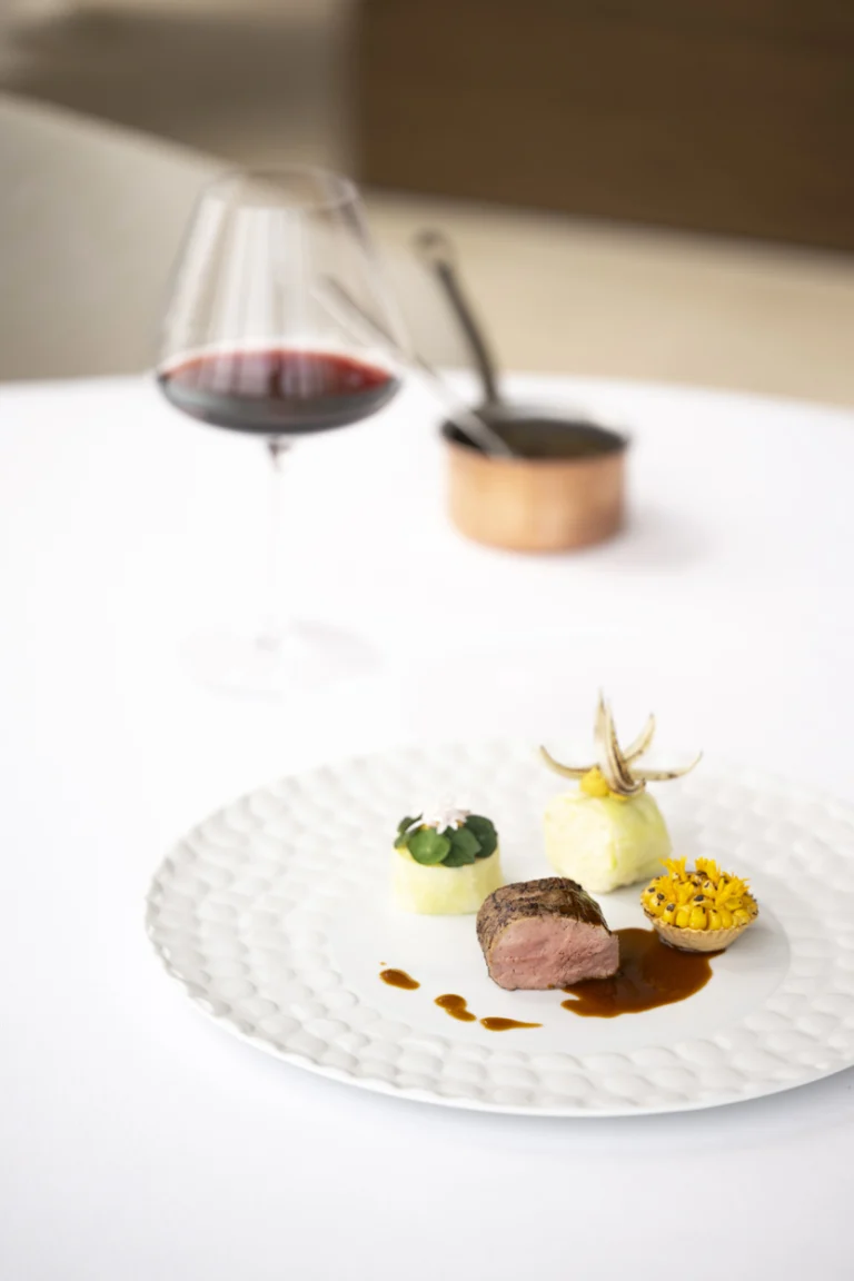A plated dish of meat with garnishes beside a glass of red wine on a white tablecloth