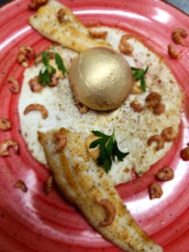 Two fish fillets with small brown shrimps and a gold-coloured sphere on a red plate