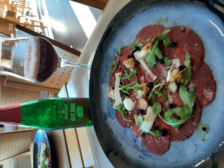 Beef carpaccio with rocket and shavings of cheese on a dark plate, with a glass and a green bottle at the edge of the frame