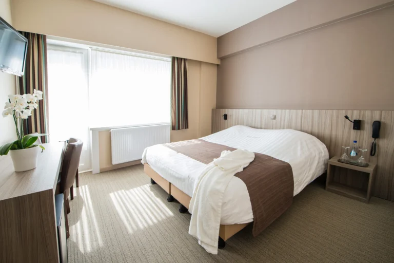 Sunlit double room with a white-and-brown bed, a wooden desk with an orchid and a large curtained window