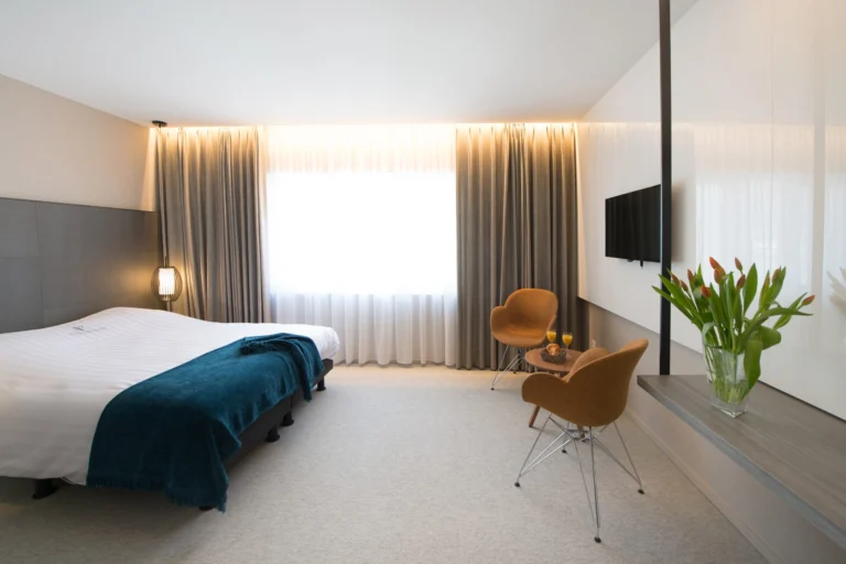Bright hotel room with a double bed and teal throw, two orange chairs, a wall-mounted TV and tulips in a vase