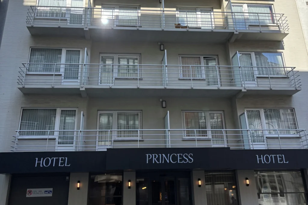 Pale facade of Hotel Princess with rows of balconies and a dark awning bearing the hotel name above the entrance