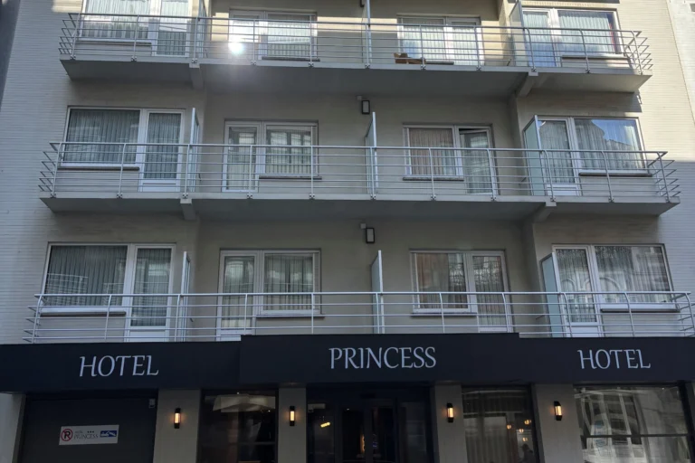 Pale facade of Hotel Princess with rows of balconies and a dark awning bearing the hotel name above the entrance