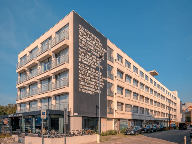 Modern five-storey Hotel Royal Astrid with balconies and a large poem on its dark corner wall, under a blue sky