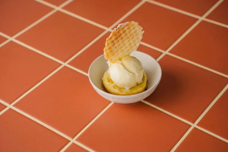 A small bowl with a scoop of ice cream on a round yellow base, topped with a thin waffle wafer, on a terracotta-tiled surface
