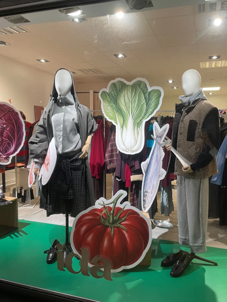 Ida's shop window with two mannequins in layered autumn outfits among oversized cut-outs of a tomato, a pak choi and a fish, with clothing rails visible inside