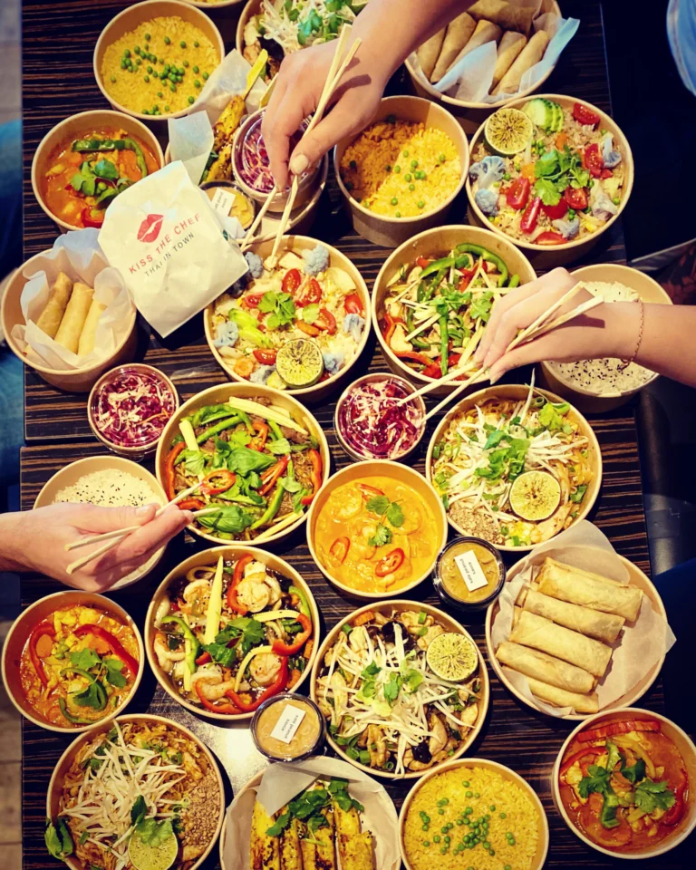 A table covered with takeaway bowls of Thai curries, stir-fries and spring rolls, with hands reaching in with chopsticks