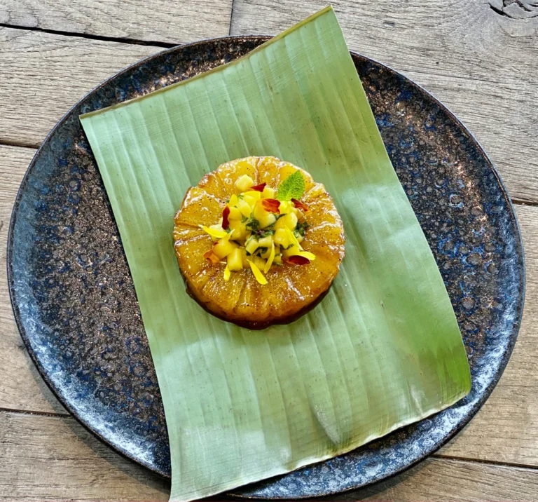 A glazed golden disc topped with diced mango and edible flowers, served on a banana leaf on a dark plate