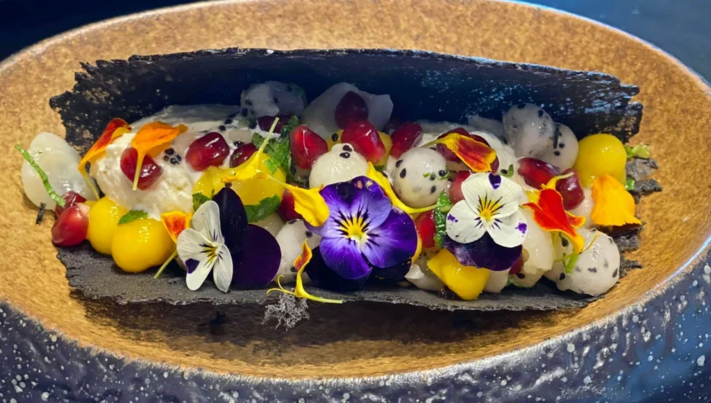 A colourful plated dish topped with edible pansies, pomegranate seeds and small pieces of fruit on a dark strip, in a speckled stoneware bowl