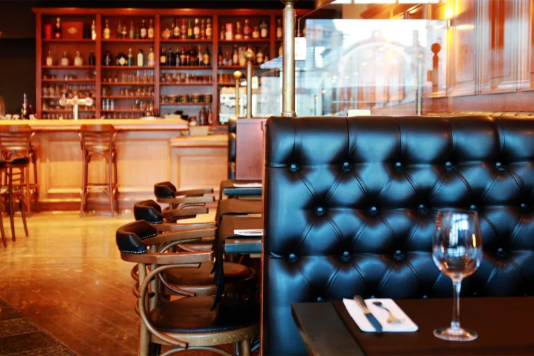 Wooden bar with shelves of bottles behind a black leather booth and a table set with a wine glass