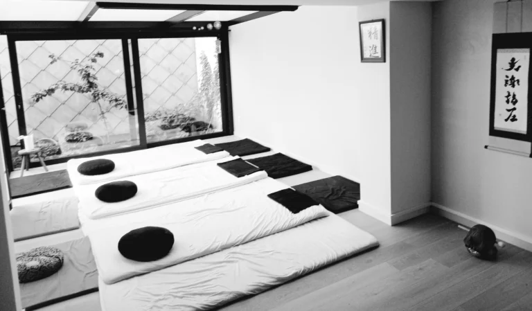 Black-and-white photo of futons and meditation cushions laid out in rows in a dojo with Japanese calligraphy on the wall