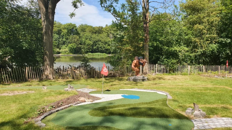 Minigolf hole with a caveman statue beside the lake, surrounded by tall trees