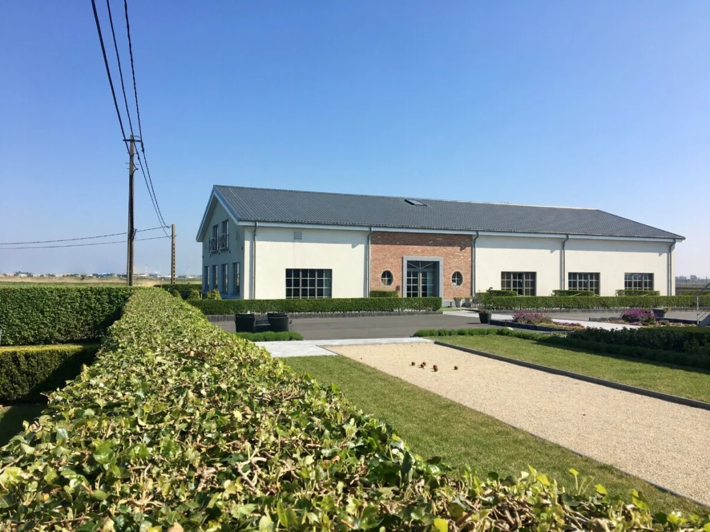 Low brick and white-rendered hotel building behind clipped hedges and a pétanque court under a blue sky