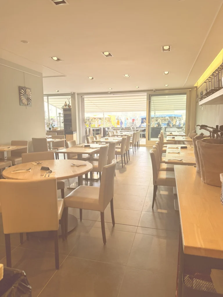 Long dining room with cream chairs and wooden tables, its glass front looking out on stalls on the quay