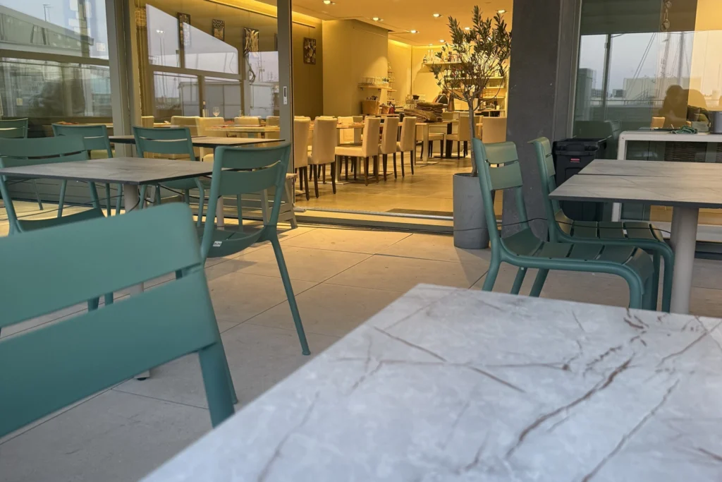 Terrace tables with marble-effect tops and green chairs in front of open glass doors to a lit dining room
