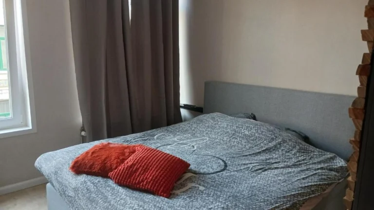 Guest room with a grey double bed, red scatter cushions and daylight from a tall window