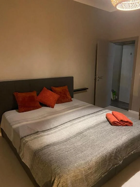 Double room with a dark headboard, grey bedding, orange cushions and a folded towel, with the bathroom door ajar