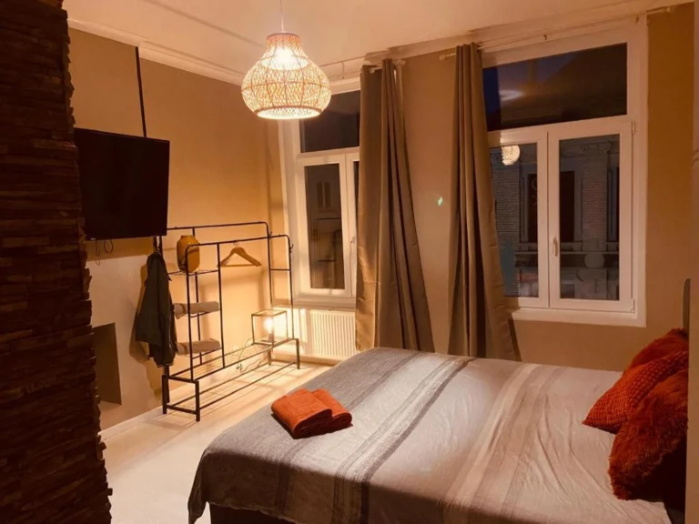 Double room at La Nonne Blanche with a warm interior, a pendant light, orange cushions on a white bed and a coat rack by the window