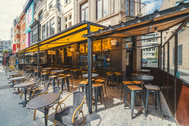 Covered terrace with a yellow awning, high tables and stools along the Langestraat in front of the bar