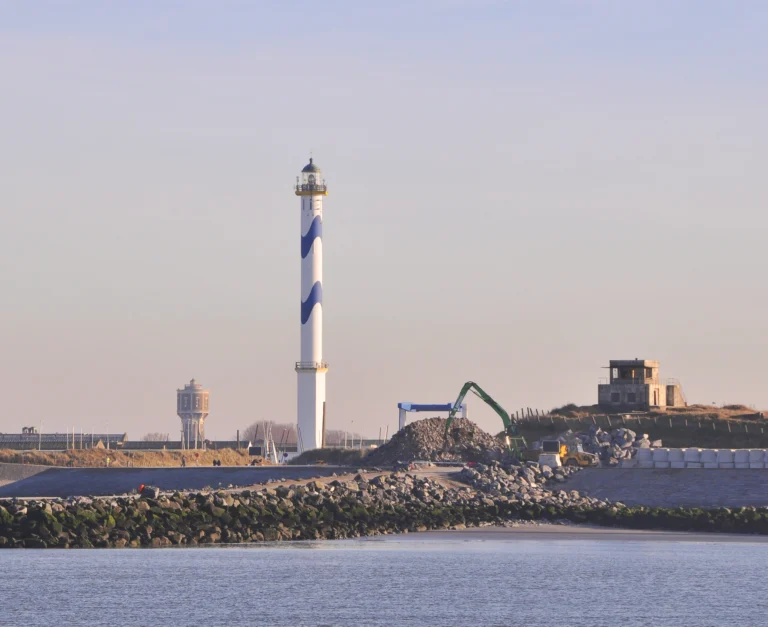The lighthouse seen across the harbour channel in soft evening light, with a stone breakwater in the foreground