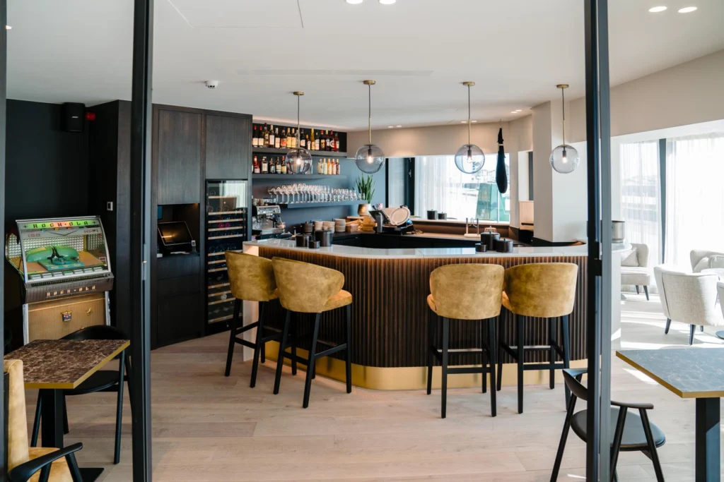Curved bar with ochre velvet bar stools, glass globe pendant lamps, a wine fridge and a vintage Rock-Ola jukebox at Le Bar du Bassin