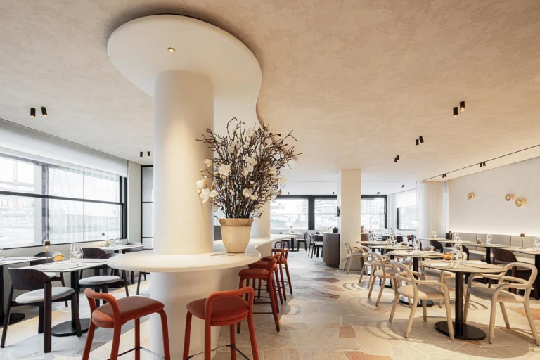 Brasserie dining room with a curved white bar column, rust-red bar stools, a stone floor and harbour-side windows