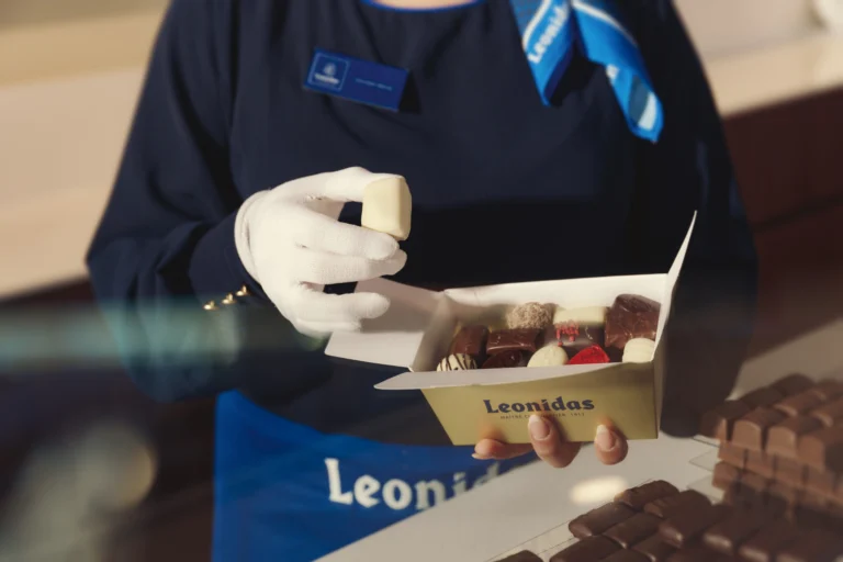 A Leonidas shop assistant in white gloves placing a white praline into an open gold ballotin already half filled with assorted chocolates