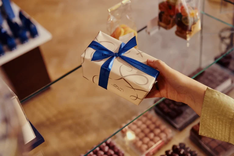 A hand holding a white and gold gift-wrapped box tied with a blue ribbon above a glass counter of chocolates