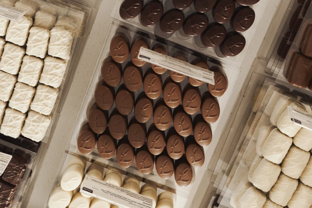 Rows of milk, dark and white chocolate pralines laid out in trays behind the glass of a Leonidas counter, each tray with a small name card